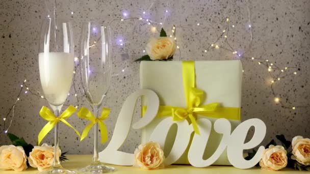 Lo champagne viene versato in un bicchiere. Due bicchieri di champagne con fiocchi gialli in piedi sul tavolo con lettere d'amore, regalo e rose, luci bokeh, biglietto di auguri per San Valentino o matrimonio - Filmati, video