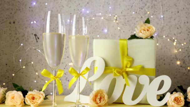Two glasses with yellow bows of champagne standing on table with love letters, gift and roses, bokeh lights, St. Valentine or wedding greeting card - Footage, Video