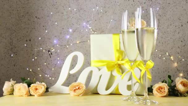 Dos copas de champán con arcos amarillos de pie en la mesa con cartas de amor, regalo y rosas, luces bokeh enrolladas de círculos, enfoque selectivo, tarjeta de felicitación de San Valentín o boda - Metraje, vídeo