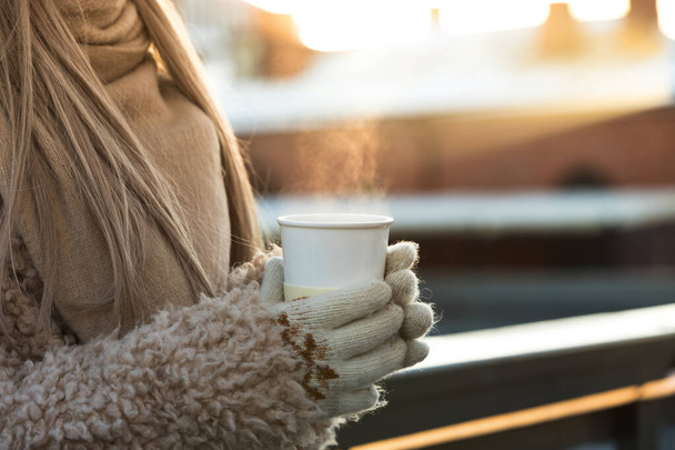 Manos femeninas en manoplas blancas que sostienen la taza blanca humeante de café o té caliente en el día soleado frío del invierno, primer plano. - Foto, Imagen