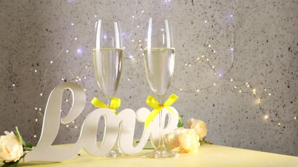 Dvě sklenice na šampaňské se žlutými luky stojící na stole s milostnými písmeny a růžemi válcovanými z kruhů, bokeh světly, sv. Valentýna nebo svatební přání - Záběry, video