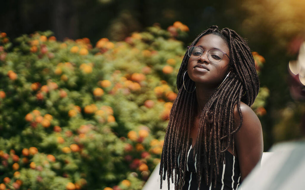 A ravishing young black female with dreadlocks and eyeglasses in the public park with a bush with flowers in the defocused background; a portrait of a dazzling African woman with dreads and in glasses - Fotografie, Obrázek