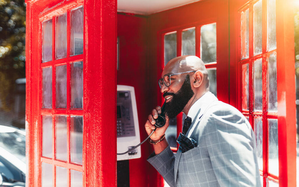 A cheerful mature bald elegant black man entrepreneur in a fashionable business costume, eyeglasses, and with a well-groomed beard is speaking on a telephone standing in a retro red phone-booth - Фото, зображення