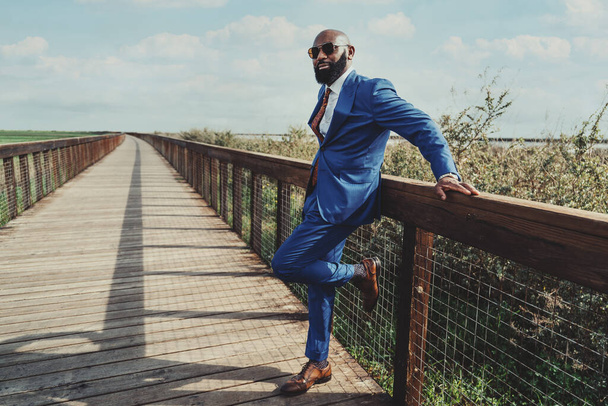 Portrait of a mature bald bearded black man in sunglasses and an elegant custom made blue suit with a necktie and leather shoes is leaning on a fence of a wooden bridge stretching into the distance - Photo, Image