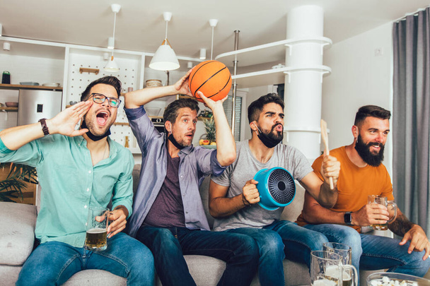 Χαρούμενοι άντρες φίλοι που φοράνε προστατευτική μάσκα με μαύρο πρόσωπο βλέποντας μπάσκετ στο σπίτι - Φωτογραφία, εικόνα