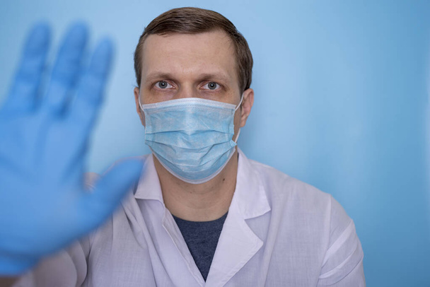 Ein männlicher Arzt in medizinischer Maske zeigt mit seiner Hand auf blauem Hintergrund einen Anschlag. Das Konzept der Medizin und des Virenschutzes. - Foto, Bild