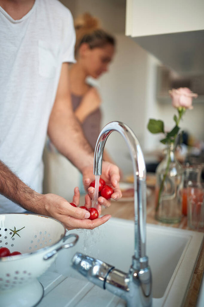 A young man washing fresh cherry tomatoes under the tap in the kitchen on a beautiful morning. Home, kitchen, salad - Foto, Imagem