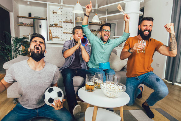 Felice amici maschi con vuvuzela indossare maschera protettiva viso nero guardando il calcio a casa - Foto, immagini