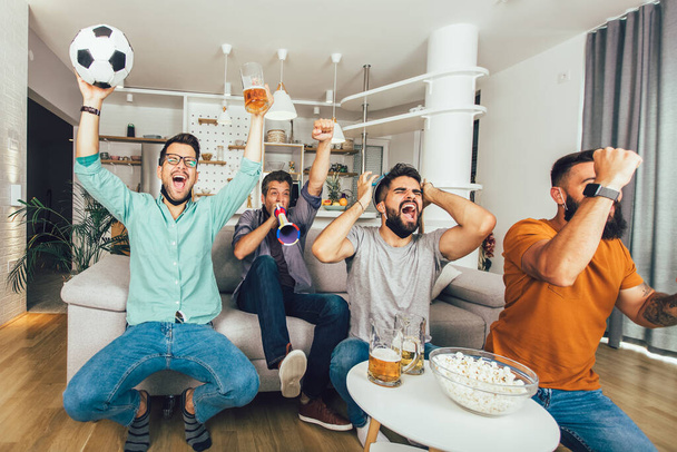 Glückliche männliche Freunde mit Vuvuzela, die eine schwarze Gesichtsschutzmaske tragen und zu Hause Fußball schauen - Foto, Bild