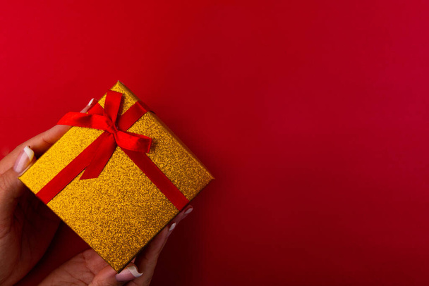 Woman's hands hold diagonally gift box wrapped in red ribbon isolated on red background - Foto, afbeelding