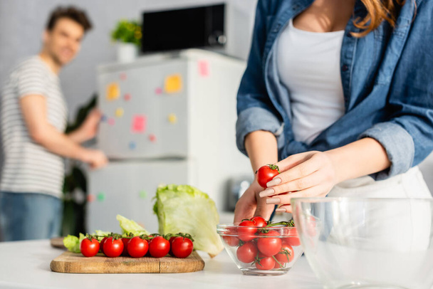 cropped view of woman holding red cherry tomato near ingredients on kitchen table and boyfriend on blurred background  - Photo, image