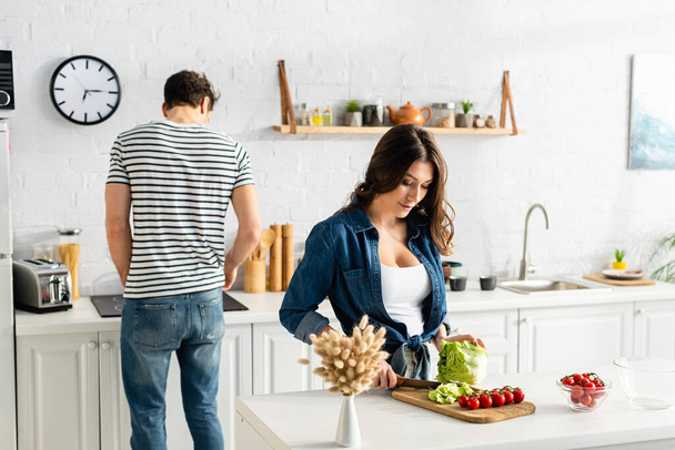woman cutting lettuce near ingredients on kitchen table and boyfriend - Фото, зображення
