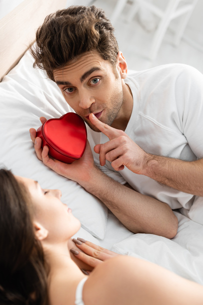 mężczyzna trzyma czerwone pudełko w kształcie serca i pokazuje znak ciszy w pobliżu śpiącej kobiety na niewyraźnym pierwszym planie  - Zdjęcie, obraz