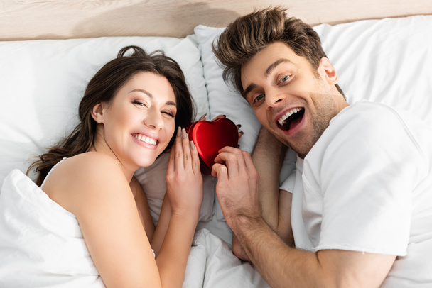 κορυφαία άποψη του ευτυχισμένου ζευγαριού που βρίσκεται στο κρεβάτι κοντά στο κόκκινο κουτί σε σχήμα καρδιάς για την ημέρα του Αγίου Βαλεντίνου - Φωτογραφία, εικόνα