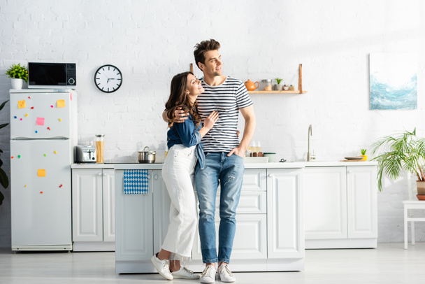 full length of happy woman hugging cheerful man standing with hand in pocket in modern kitchen  - 写真・画像