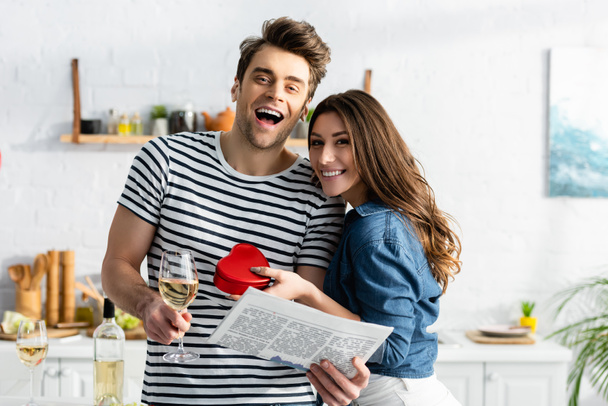 man holding glass of wine and newspaper near cheerful girlfriend with heart-shaped gift box - Foto, afbeelding