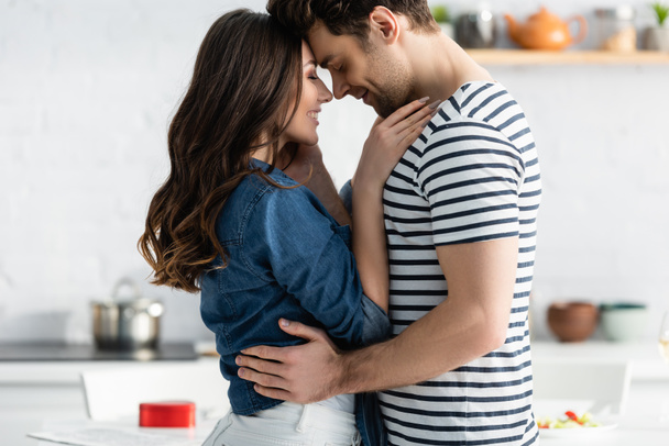 vista lateral de pareja feliz con los ojos cerrados abrazándose en la cocina  - Foto, Imagen