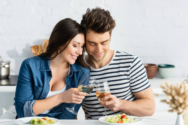 happy couple toasting glasses of wine near plates with salad in kitchen  - Фото, зображення