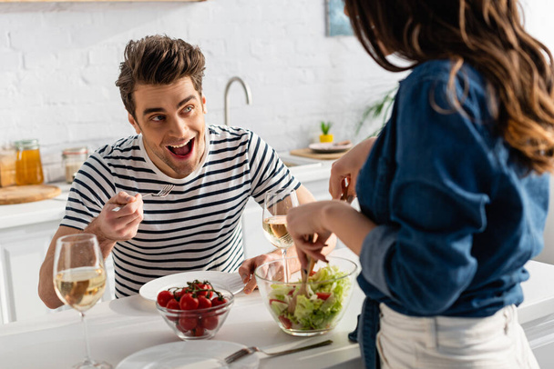 homem espantado olhando para a mulher servindo salada na cozinha em primeiro plano borrado  - Foto, Imagem