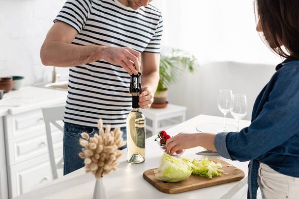 женщина режет салат возле бойфренда открывая бутылку вина - Фото, изображение