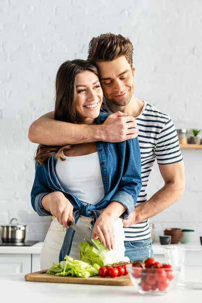 happy man hugging woman preparing salad in kitchen  - Valokuva, kuva