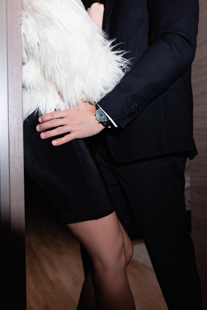 Gestroopt uitzicht op de man in pak aanraken vriendin in jurk en pluizig jasje in hotelkamer  - Foto, afbeelding