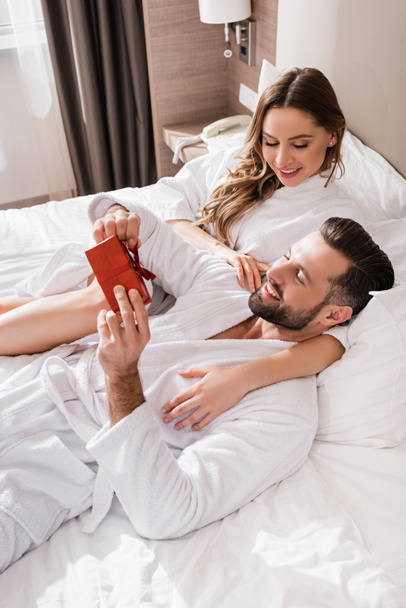 Man in bathrobe holding present near smiling girlfriend on hotel bed  - Фото, изображение