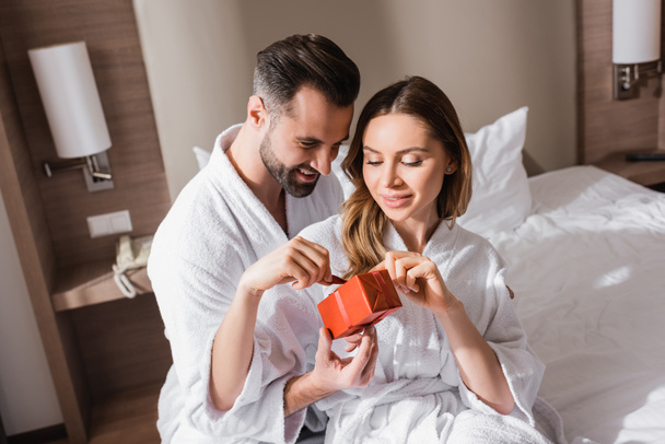 Улыбающаяся пара в халатах, держащая подарок на кровати отеля  - Фото, изображение