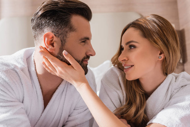 Smiling woman in bathrobe touching face of boyfriend in hotel room  - Photo, Image