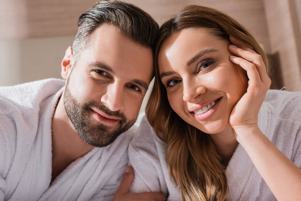 Positive couple in white bathrobes looking at camera in hotel  - Φωτογραφία, εικόνα