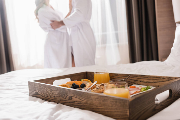 Cropped view of delicious breakfast on tray on bed and couple in bathrobes on blurred background in hotel  - Photo, Image