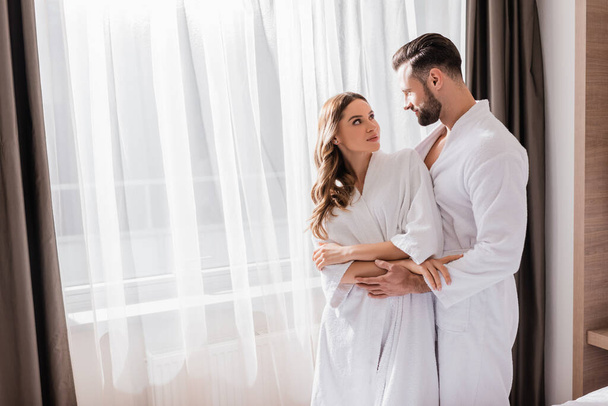 Paar in weißen Bademänteln umarmt sich am Fenster im Hotelzimmer  - Foto, Bild