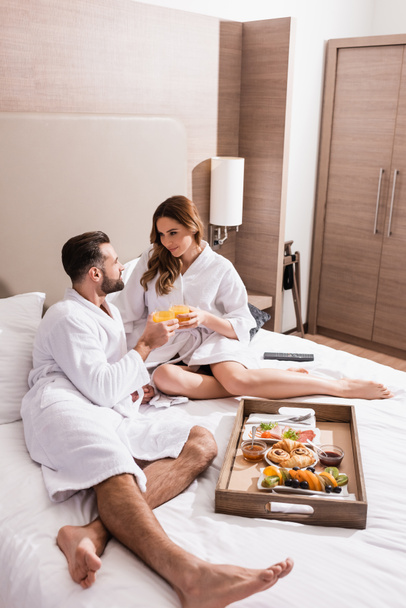 Couple in bathrobes holding glasses of orange juice near breakfast on tray on hotel bed  - Photo, Image