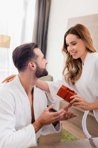 Lächelnde Frau umarmt Freund im Bademantel und hält Geschenk im Hotelzimmer  - Foto, Bild