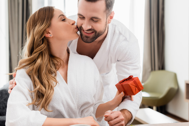 Mujer en albornoz besando novio y sosteniendo caja de regalo en habitación de hotel  - Foto, Imagen