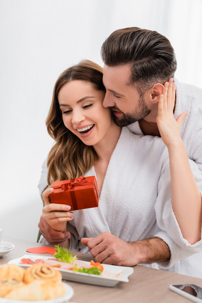 Cheerful woman in bathrobe touching boyfriend while holding gift box near breakfast on blurred foreground in hotel  - Фото, изображение