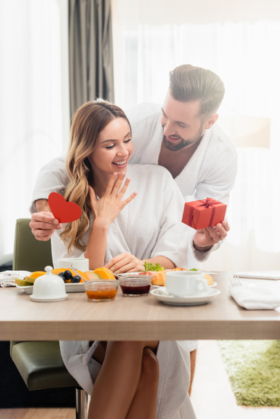 Uomo sorridente che tiene presente e cuore di carta vicino alla fidanzata in accappatoio e colazione in primo piano sfocato in hotel  - Foto, immagini