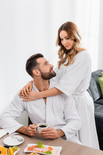 Mujer en albornoz abrazando novio con taza de café cerca del desayuno en primer plano borroso en el hotel  - Foto, imagen