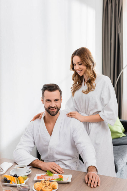Cheerful woman in bathrobe hugging boyfriend near breakfast with coffee on table in hotel room  - Foto, afbeelding