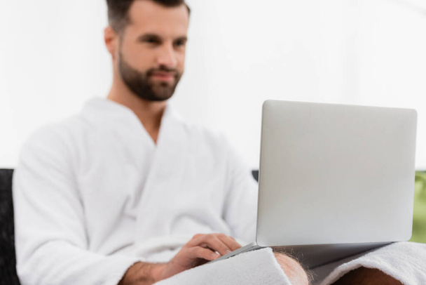 Laptop on laps of man in bathrobe on blurred background in hotel room  - Fotoğraf, Görsel