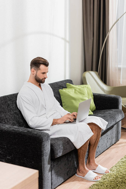 Terlik ve bornoz giyen bir tele-işçi oteldeki kanepede dizüstü bilgisayar kullanıyor.  - Fotoğraf, Görsel