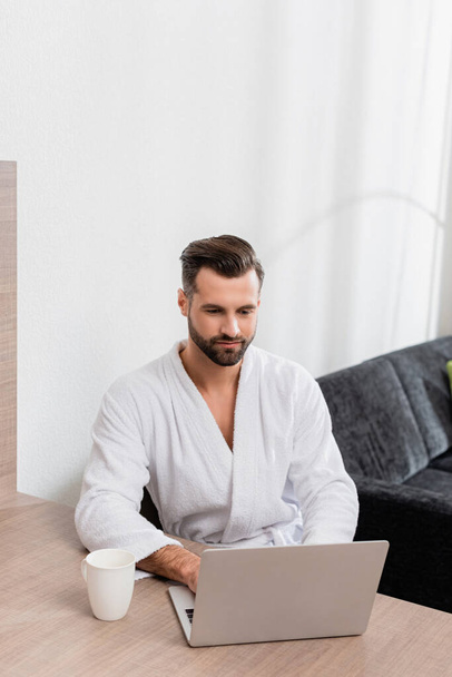 Man in white bathrobe using laptop near cup on table in hotel room  - Fotoğraf, Görsel