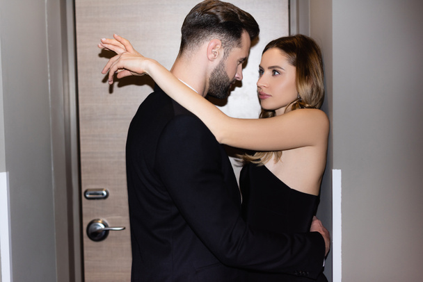Man in formal wear hugging seductive girlfriend in dress in hotel room  - Photo, Image