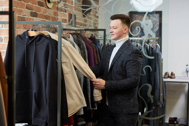 Ein junger, stylischer Mann im Anzug wählt in einem Bekleidungsgeschäft ein Hemd oder einen Kapuzenpullover. Ein männlicher Kunde in einer Boutique. - Foto, Bild