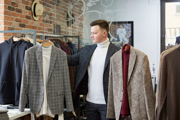 Ένας νεαρός άνδρας με ένα λευκό πουλόβερ ζιβάγκο και ένα κοστούμι διαλέγει ανάμεσα σε δύο κοστούμια σε κρεμάστρες σε ένα κατάστημα ρούχων. Ένας άντρας πελάτης σε μια μπουτίκ. - Φωτογραφία, εικόνα