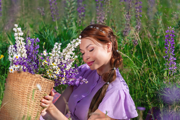 Portrét mladé krásné ženy s kyticí květin v košíku v létě příroda sedí na trávě. Pěkná vintage dívka v šeříku šaty se usmívá na slunné lesní louce. - Fotografie, Obrázek