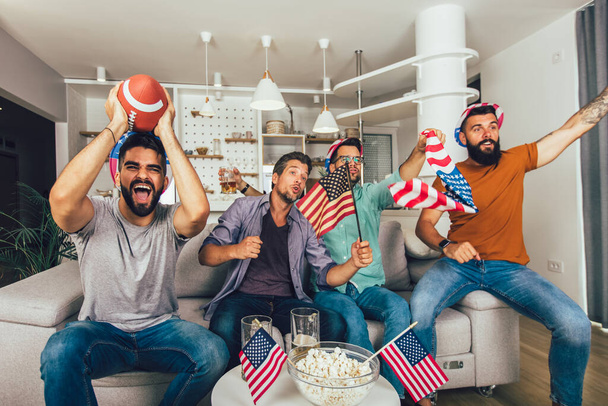 Ενθουσιασμένοι άνδρες φίλοι ζητωκραυγάζουν βλέποντας αμερικανικό ποδοσφαιρικό αγώνα στον καναπέ στο σπίτι. - Φωτογραφία, εικόνα