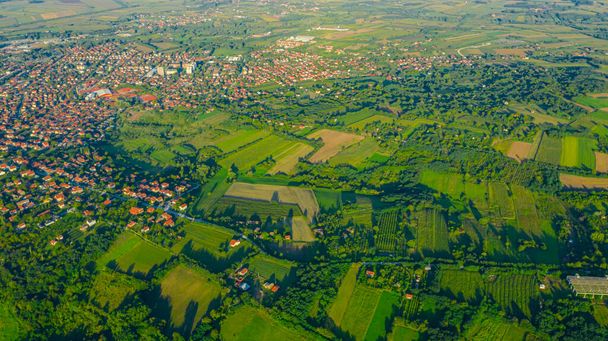 Shot above over green hilly landscape, several cultivated plots among forest trees and city in the distance. - Photo, Image