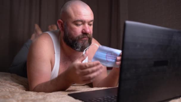 ベッドの上の自宅でTシャツを着た髭の男は、心臓が描かれた医療用保護マスクを着用しています。オンラインデートの概念。バレンタインデー - 映像、動画