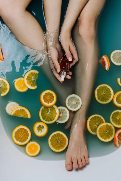 Faceless shot of woman in a bathroom filled with blue water and various cut citrus fruits holding serum in her hands - Foto, immagini